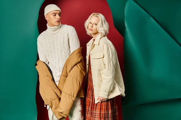 Hombre con estilo en gorro y chaqueta hinchable mirando a la mujer rubia en rojo con fondo turquesa - foto de stock