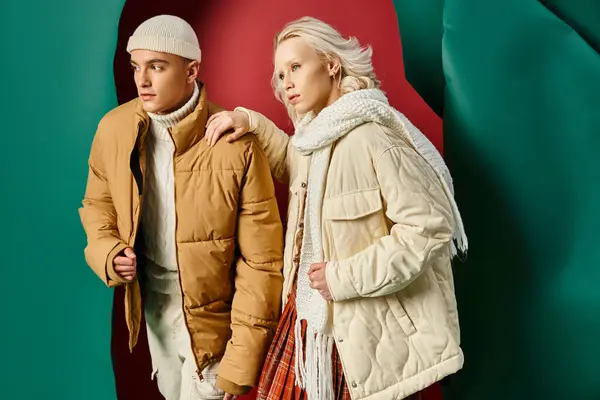 Elegante uomo in beanie e giacca di puffer in posa con donna bionda sul rosso con sfondo turchese — Foto stock