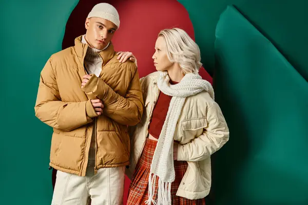 Junge blonde Frau im karierten Rock schaut Mann in Winterkleidung auf rotem mit türkisfarbenem Hintergrund an — Stockfoto