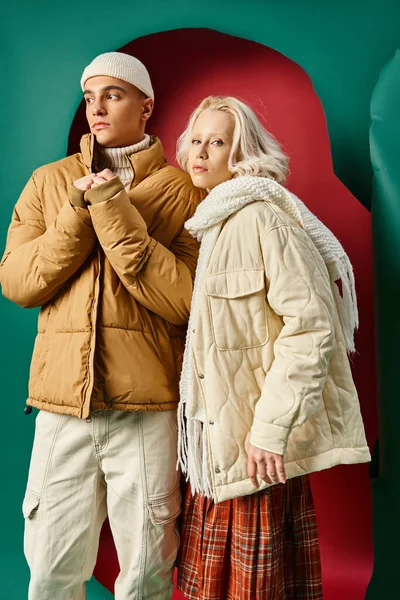 Joven mujer rubia en falda a cuadros posando con el hombre en chaqueta de invierno en rojo con fondo turquesa - foto de stock