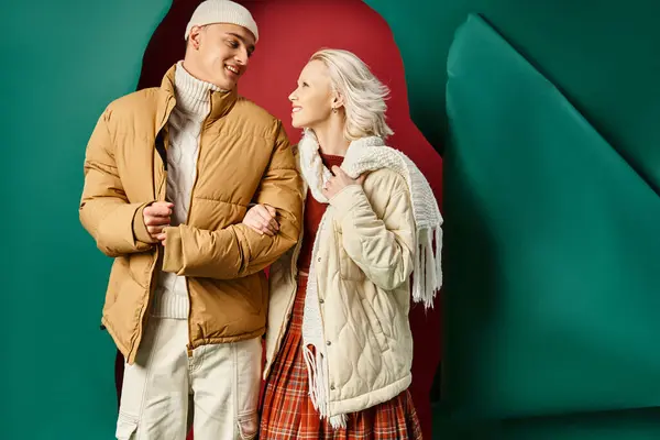 Casal alegre em jaquetas de inverno elegantes sorrindo e posando juntos no vermelho com pano de fundo turquesa — Fotografia de Stock