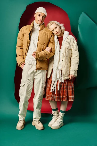 Longueur totale de jeune couple en vestes d'hiver posant ensemble sur fond rouge avec fond turquoise — Photo de stock