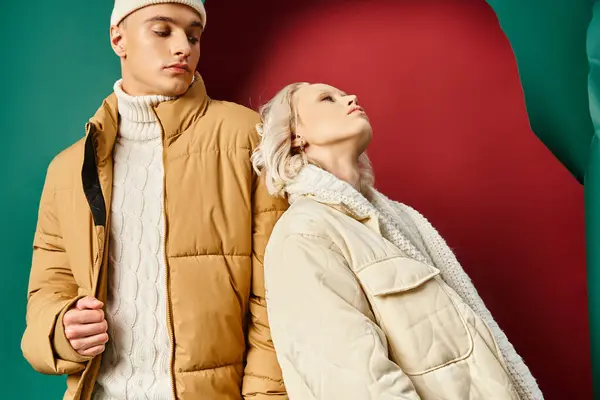 Blonde woman in winter jacket leaning on handsome man in beanie on red with turquoise backdrop — Stock Photo