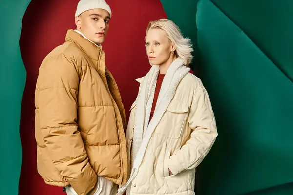 Пара в модных зимних куртках позирует с руками в карманах на красном с бирюзовым фоном — стоковое фото