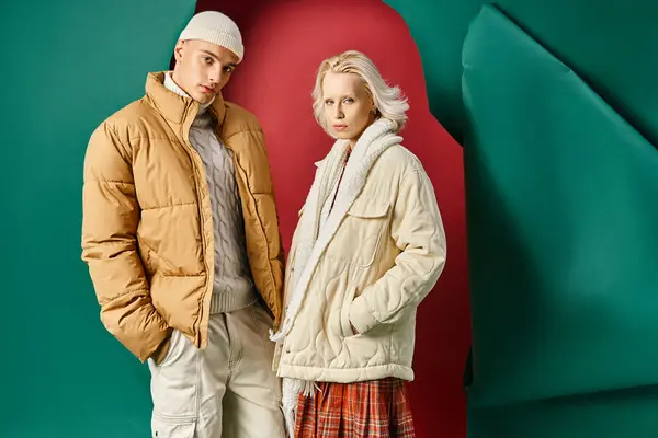 Блондинка и мужчина в зимних куртках позируют с руками в карманах на красном с бирюзовым фоном — стоковое фото