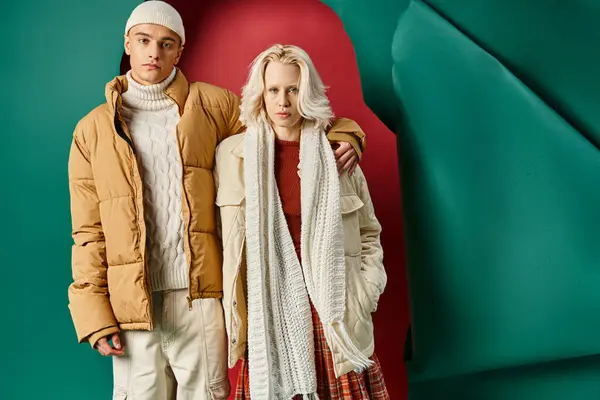 Man in beanie hugging blonde woman and standing in winter outerwear on red with turquoise backdrop — Stock Photo
