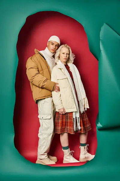 Longitud completa de hombre y mujer con estilo en chaquetas de invierno posando cerca de color turquesa desgarrado y fondo rojo - foto de stock