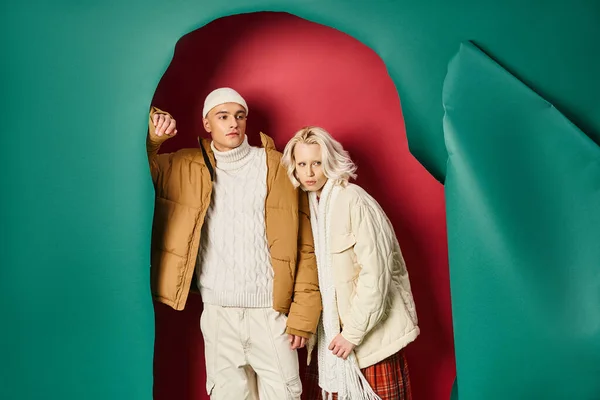 Schöner Mann in Mütze und Winterjacke neben stilvoller Frau auf zerrissenem türkis-rotem Hintergrund — Stockfoto