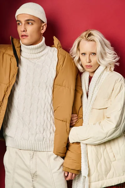 Jeune femme blonde en blanc hiver vêtements d'extérieur tenant la main de l'homme en bonnet chapeau sur fond rouge — Photo de stock