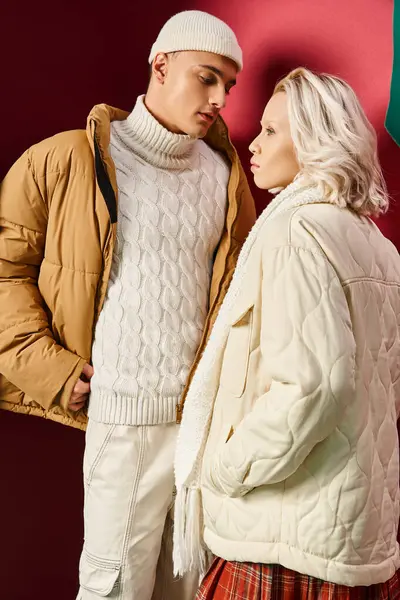 Блондинка в белой зимней куртке позирует рядом со стильным мужчиной в шапочке на красном фоне — стоковое фото