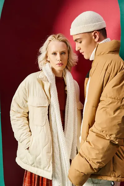Blondine in Winterjacke posiert mit Händen in Taschen neben stilvollem Mann auf rotem Hintergrund — Stockfoto