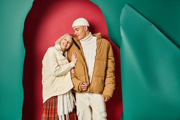 Mujer complacida en chaqueta de invierno abrazando novio feliz cerca de desgarrado turquesa y fondo rojo - foto de stock
