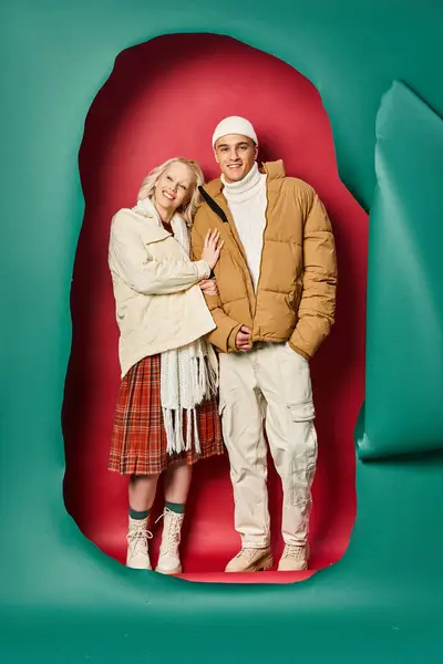 Glückliche Frau in Winterjacke umarmt ihren fröhlichen Freund in der Nähe von zerrissenem Türkis und rotem Hintergrund — Stockfoto