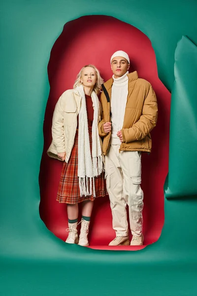 Blonde woman in winter jacket and plaid skirt posing with man near torn turquoise and red backdrop — Stock Photo