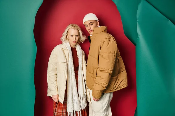 Young woman in winter outerwear and plaid skirt posing with man near torn turquoise and red backdrop — Stock Photo