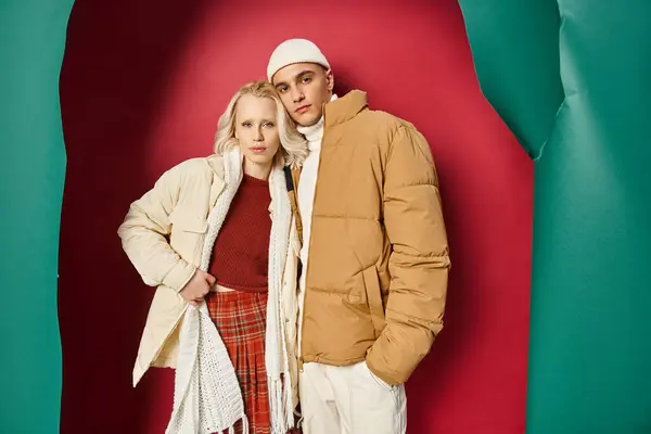 Atractiva mujer y hombre en ropa de abrigo de invierno posando juntos cerca de color turquesa desgarrado y fondo rojo - foto de stock