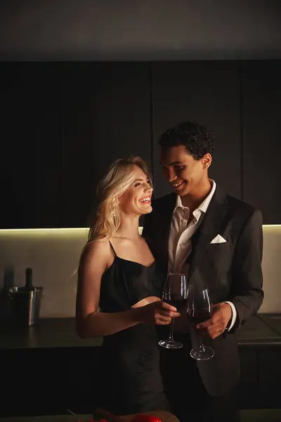 Fröhliches junges multikulturelles Paar in eleganter Kleidung, das einander genießt und Weingläser in der Hand hält — Stockfoto