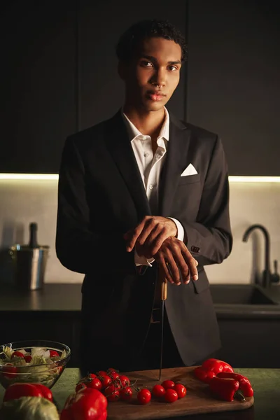 Joven guapo en elegante traje negro posando en su cocina y mirando directamente a la cámara - foto de stock