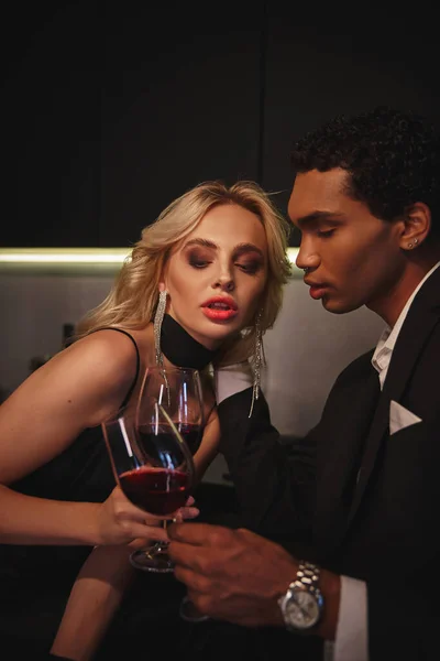 Young sensual multicultural couple in evening attires clinking glasses of red wine in kitchen — Stock Photo