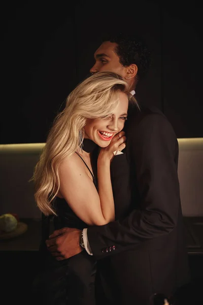 Alegre hermosa mujer con cabello rubio sonriendo y abrazando a su novio afroamericano - foto de stock