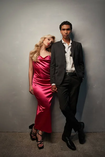 Atractiva pareja multirracial amorosa en trajes de noche tomados de la mano y posando sobre fondo gris - foto de stock