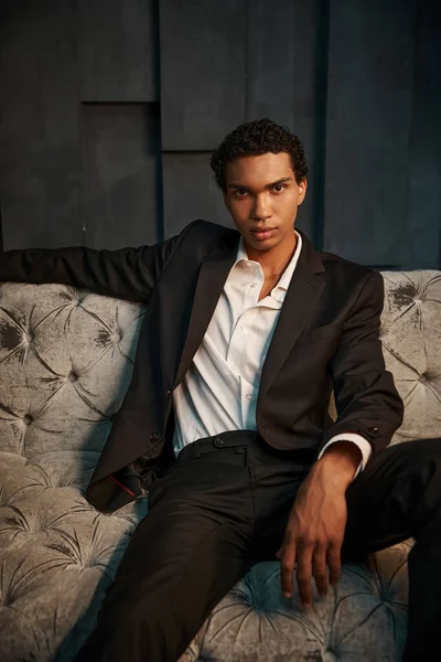 Handsome relaxed african american male model in chic suit sitting on sofa and looking at camera — Stock Photo