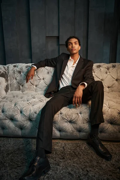 Handsome young african american man in elegant suit sitting on comfy sofa and looking at camera — Stock Photo