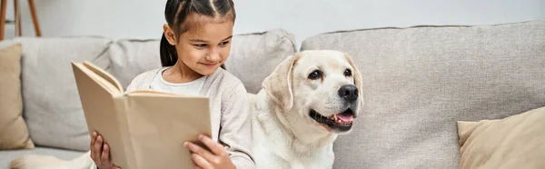Ragazza felice seduta sul divano e libro di lettura vicino labrador in soggiorno moderno, banner compagno — Foto stock