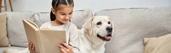 Happy girl sitting on sofa and reading book near labrador in modern living room, companion banner — Stock Photo