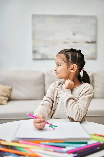 Distracted cute elementary age girl drawing with color pencil on paper in modern apartment — Stock Photo