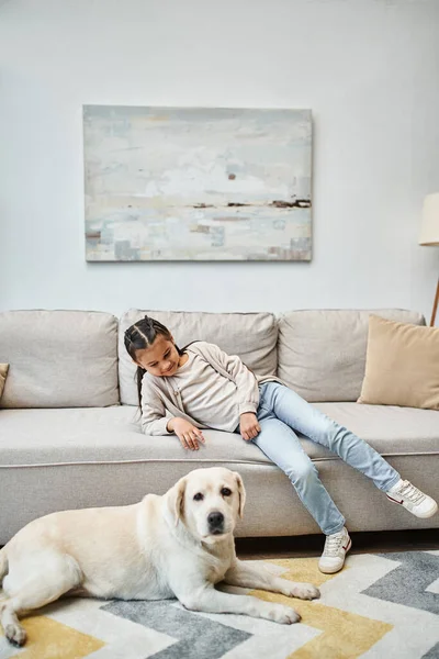 Menina bonito sorrindo e sentado no sofá enquanto olha para o labrador na sala de estar moderna, criança e cão — Fotografia de Stock