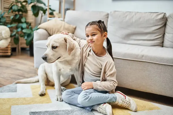 Cute girl smiling and sitting near sofa while stroking labrador in modern living room, kid and dog — Stock Photo