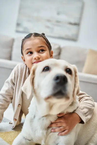 Счастливая симпатичная девочка, сидящая на ковре и гладящая собаку в современной гостиной, малыш и лабрадор — стоковое фото