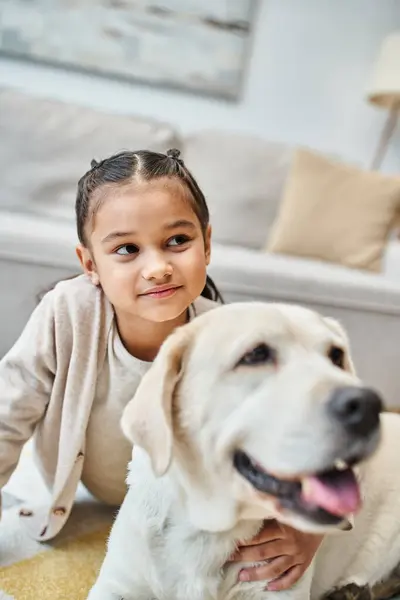 Feliz linda chica sentada en la alfombra y acariciando perro en la sala de estar moderna, niño y labrador — Stock Photo