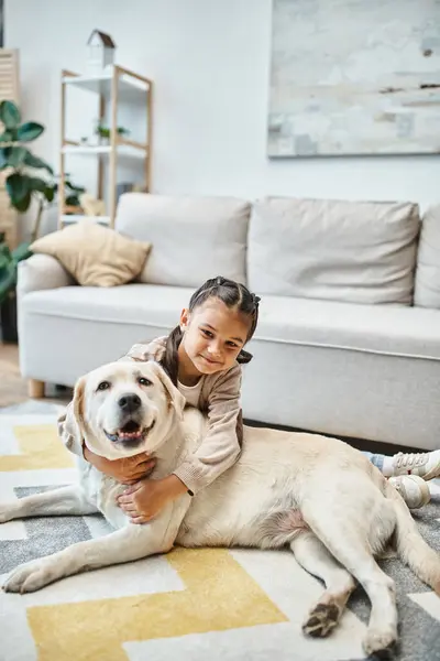 Cheerful girl in casual attire smiling and stroking dog in modern living room, child and labrador — Stock Photo