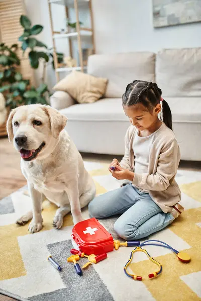 Cheerful kid in casual attire playing doctor with labrador in modern living room, toy first aid kit — Stock Photo