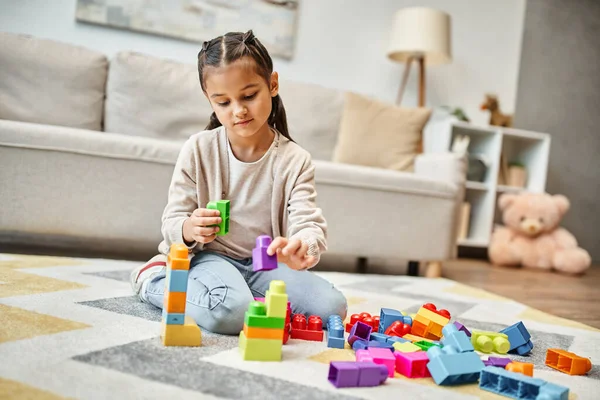 Linda menina brincando com blocos de brinquedo coloridos no tapete na sala de estar, construindo jogo de torre — Fotografia de Stock