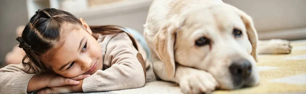 Mignonne fille d'âge élémentaire en tenue décontractée couché sur le tapis avec labrador dans le salon moderne, bannière — Photo de stock
