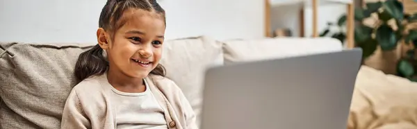 Happy elementary age girl sitting on sofa and using laptop in modern living room, e-learning banner — Stock Photo