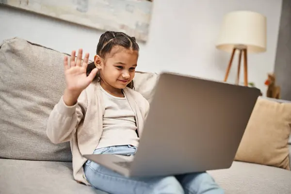 Chica de edad elemental feliz sentado en el sofá y agitando la mano en el ordenador portátil en la sala de estar, lección en línea - foto de stock