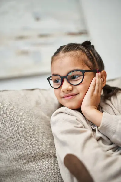 Menina idade elementar feliz em traje casual vestindo óculos e olhando para longe na sala de estar — Fotografia de Stock