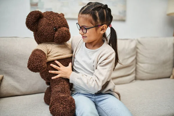 Happy girl in casual wear and eyeglasses hugging soft teddy bear and sitting on sofa in living room — Stock Photo