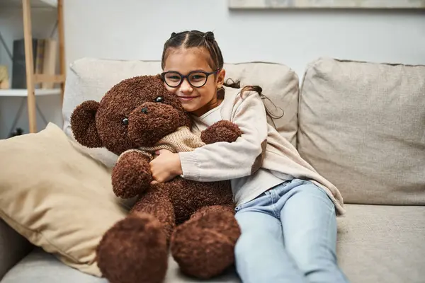 Felice ragazza in abbigliamento casual e occhiali abbracciando morbido orsacchiotto e seduto sul divano in soggiorno — Foto stock