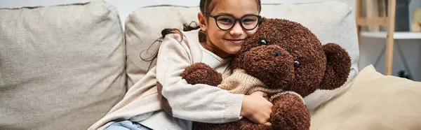 Felice ragazza in abbigliamento casual e occhiali abbracciando orsacchiotto sul divano in soggiorno, banner — Foto stock