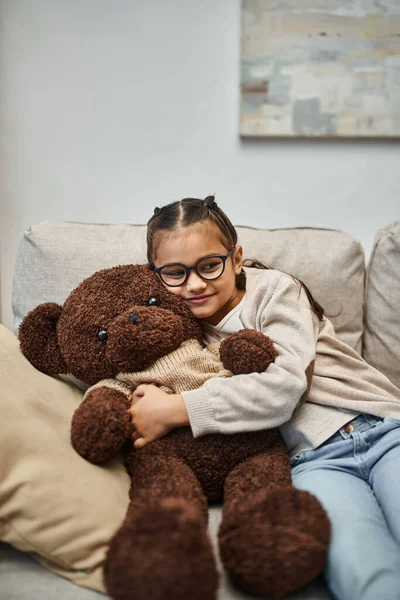 Bambino felice in abbigliamento casual e occhiali da vista abbracciando morbido orsacchiotto e seduto sul divano in soggiorno — Foto stock