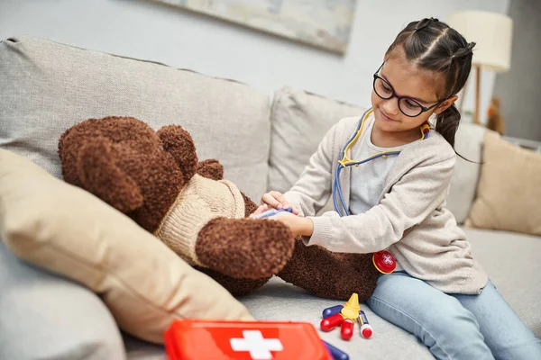 Enfant heureux en tenue décontractée et lunettes jouant médecin avec ours en peluche sur canapé dans le salon — Photo de stock