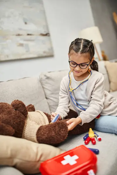 Glückliches Kind in Freizeitkleidung und Brille spielt Arzt mit Teddybär auf Sofa im Wohnzimmer — Stockfoto