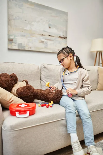 Happy kid in casual wear and eyeglasses playing doctor with soft teddy bear on sofa in living room — Stock Photo