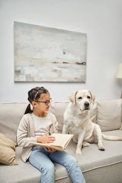 Cute girl in casual wear and eyeglasses reading book near labrador dog on sofa in living room — Stock Photo