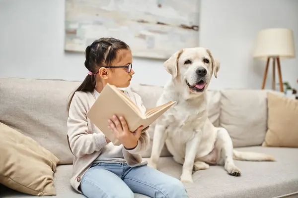 Розумна дівчина в повсякденному одязі та окулярах читає книгу для лабрадора собаки на дивані у вітальні — стокове фото